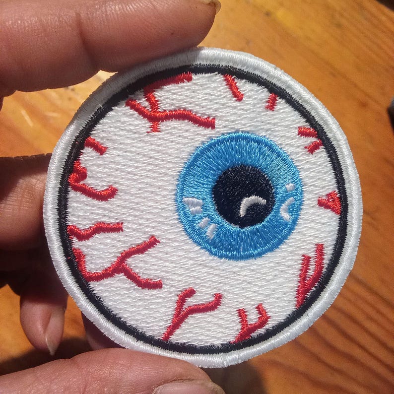 May include: Circular embroidered patch with an eyeball design. The eye has a bright blue iris, a black pupil, and red veins on a white background. The patch has a black border, suitable for application on clothing or accessories.