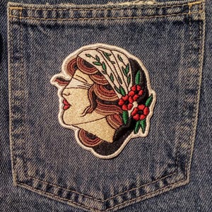 Może przedstawiać: Haftowana naszywka przedstawiająca profil kobiety z opaską na oczach, ozdobiona chustą i czerwonymi kwiatami. Naszywka umieszczona jest na kieszeni dżinsowej, podkreślając wz&oacute;r i fakturę.