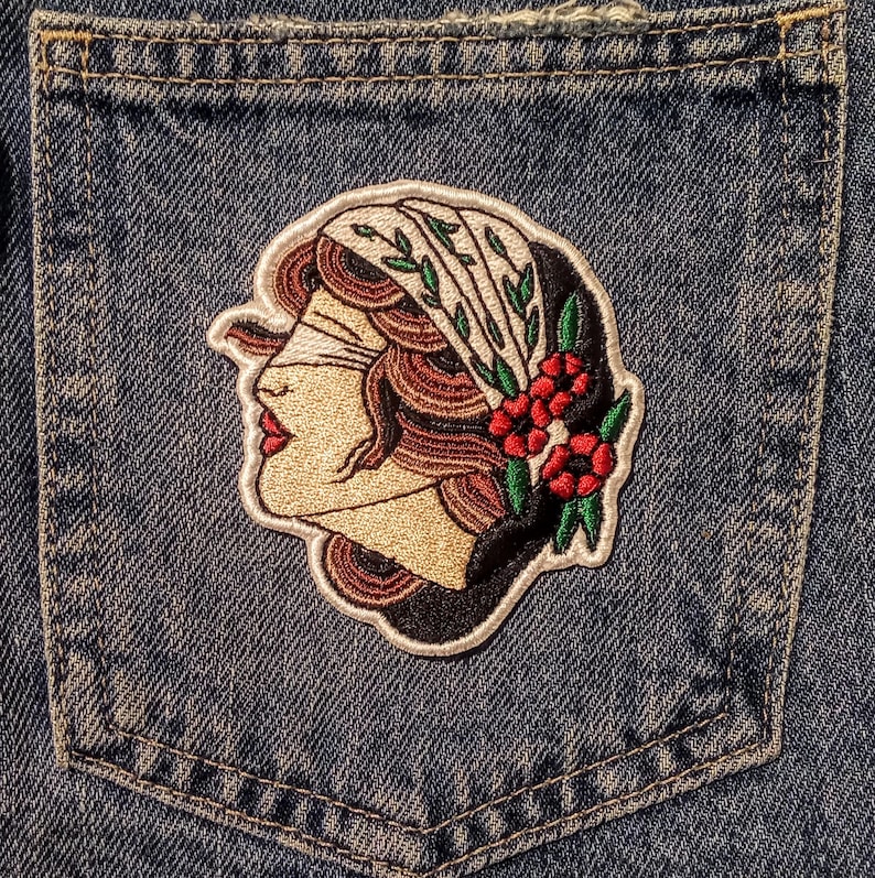 Może przedstawiać: Haftowana naszywka przedstawiająca profil kobiety z opaską na oczach, ozdobiona chustą i czerwonymi kwiatami. Naszywka umieszczona jest na kieszeni dżinsowej, podkreślając wz&oacute;r i fakturę.