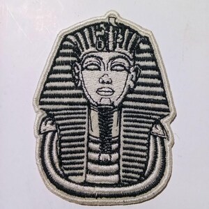 Könnte beinhalten: Gestickter Aufnäher mit einer stilisierten Darstellung der Totenmaske des Pharaos Tutanchamun. Der Aufnäher ist überwiegend schwarz-weiß, mit aufwendigen Details und einem cremefarbenen Rand. Das Design zeigt die ikonische Kopfbedeckung und die Gesichtszüge des Pharaos.