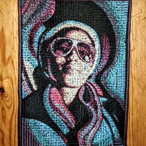 Può includere: Un'opera d'arte tessile rettangolare con un ritratto di una persona con occhiali da sole e un cappello. L'opera d'arte utilizza una varietà di colori, tra cui nero, blu, rosa e bianco, creando un aspetto strutturato e tessuto. Il pezzo è incorniciato con un bordo viola scuro.