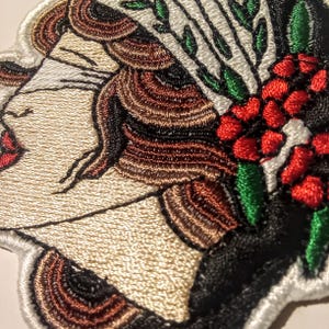 Może przedstawiać: Haftowana naszywka przedstawiająca profil kobiety z zamkniętymi oczami, czerwonymi ustami i brązowymi włosami. Projekt zawiera kompozycję kwiatową z czerwonymi jagodami i zielonymi liśćmi na czarnym tle. Naszywka ma białą obw&oacute;dkę.