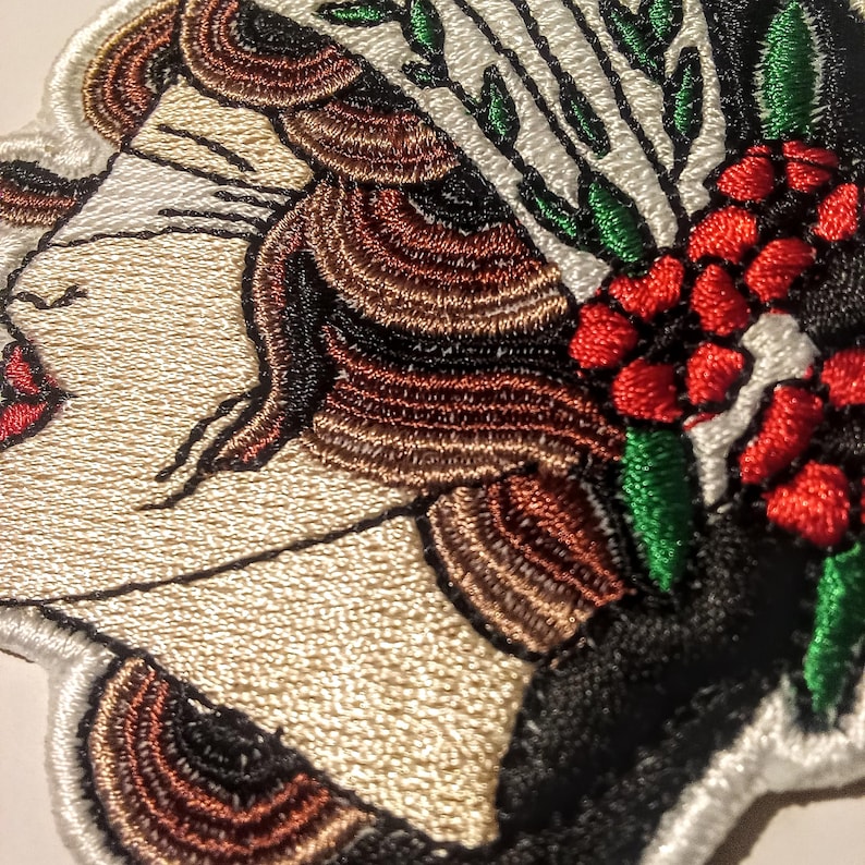 Może przedstawiać: Haftowana naszywka przedstawiająca profil kobiety z zamkniętymi oczami, czerwonymi ustami i brązowymi włosami. Projekt zawiera kompozycję kwiatową z czerwonymi jagodami i zielonymi liśćmi na czarnym tle. Naszywka ma białą obw&oacute;dkę.