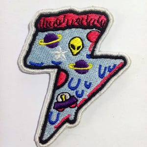 Op de afbeelding: Geborduurde patch in de vorm van een bliksemschicht, met een lichtblauwe achtergrond en een ruimtethema. Het ontwerp bevat planeten, een alien en een auto. De patch is omlijnd in zwart en wit, met rode en roze accenten. De tekst is onleesbaar.