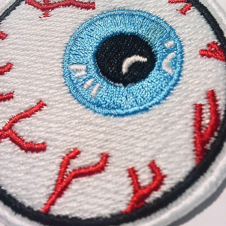 May include: Embroidered patch with an eye design. The eye has a blue iris, a black pupil, and red veins on a white background. The patch is edged in black.
