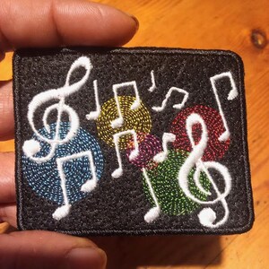 May include: Embroidered patch featuring musical notes and treble clefs in white against a black background. Colorful circles in blue, yellow, red, purple, and green are behind the musical symbols. The patch is rectangular with a black border.