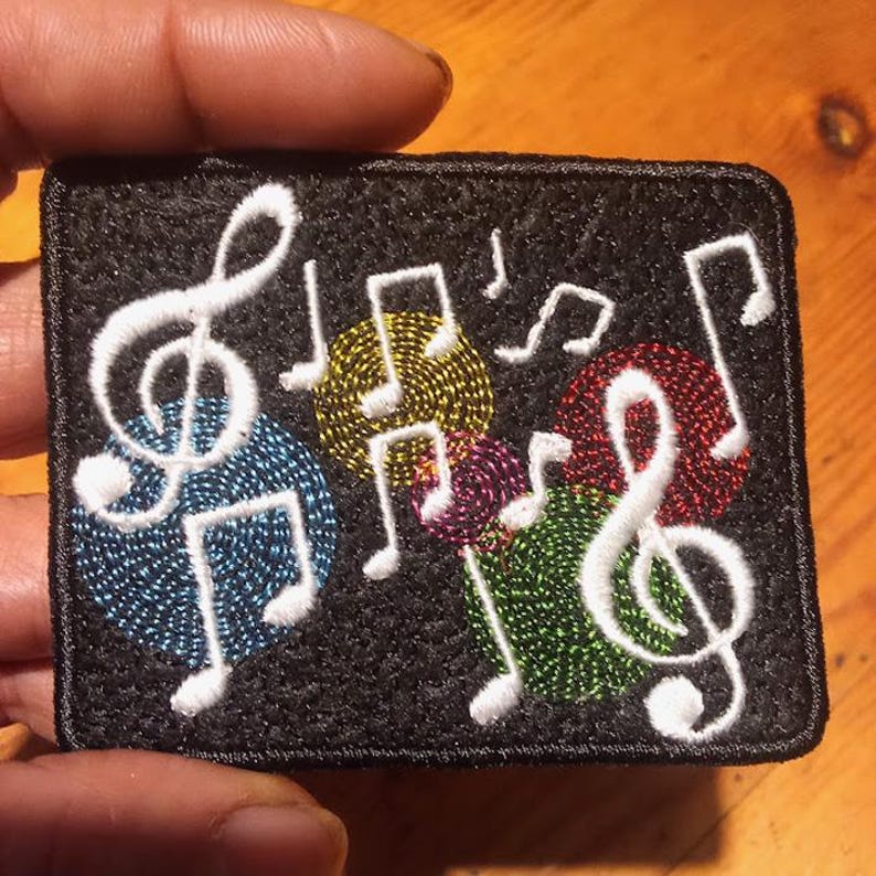 May include: Embroidered patch featuring musical notes and treble clefs in white against a black background. Colorful circles in blue, yellow, red, purple, and green are behind the musical symbols. The patch is rectangular with a black border.