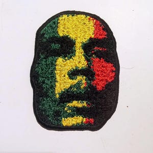 Puede incluir: Parche bordado que representa un rostro estilizado con colores verde, amarillo, rojo y negro. El parche tiene un aspecto texturizado y un borde negro. El diseño recuerda a un icono cultural.