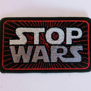 Può includere: Una toppa rettangolare ricamata con sfondo nero e bordo rosso. Il testo STOP WARS è cucito in un carattere grigio argento al centro. Linee rosse radianti si estendono dal centro dietro il testo, creando un effetto a raggiera.