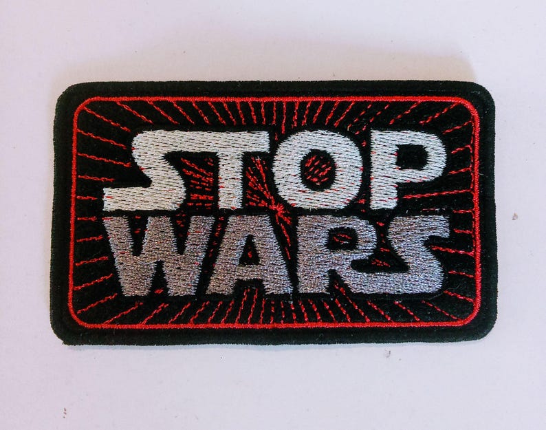 Puede incluir: Un parche rectangular bordado con fondo negro y borde rojo. El texto STOP WARS est&aacute; cosido en una fuente gris plateado en el centro. L&iacute;neas rojas radiantes se extienden desde el centro detr&aacute;s del texto, creando un efecto de rayos solares.