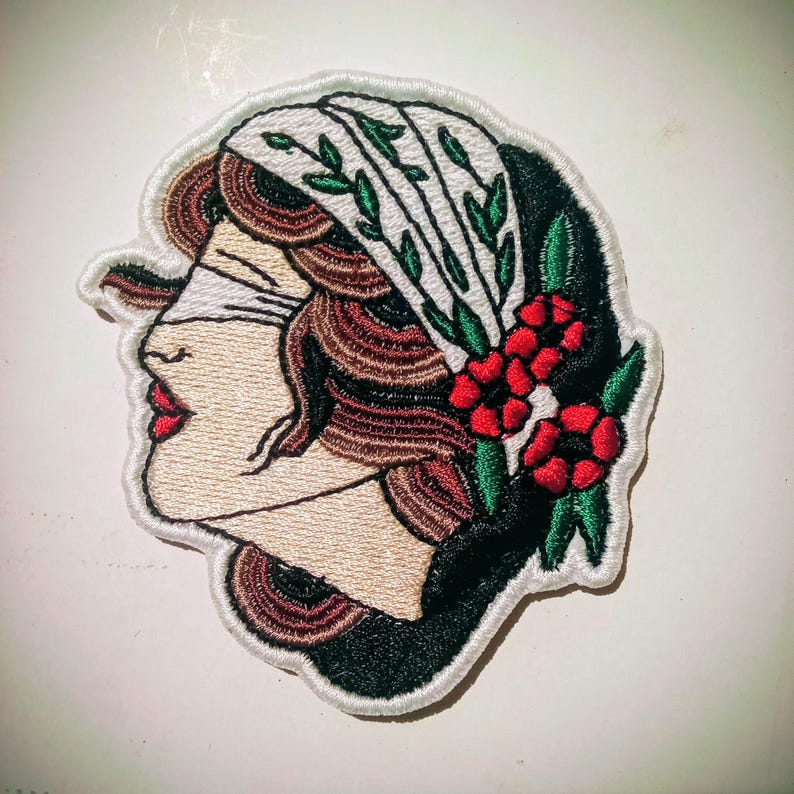 Może przedstawiać: Haftowana naszywka przedstawiająca profil kobiety z przepaską na oczach. Projekt obejmuje białe nakrycie głowy z zielonymi liśćmi, czerwonymi kwiatami i brązowymi włosami. Naszywka ma białą obw&oacute;dkę.