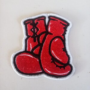 Può includere: Una toppa ricamata raffigurante un paio di guantoni da boxe rosso vivo con contorni e dettagli neri. I guantoni sono posizionati fianco a fianco, con uno che si sovrappone leggermente all'altro, rifiniti con un bordo cucito di colore bianco.