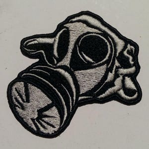 Op de afbeelding: Geborduurde patch met een gasmaskerontwerp. De patch is voornamelijk zwart met grijze details, die de contouren en kenmerken van het masker benadrukken. De patch heeft een zwarte rand en is waarschijnlijk bedoeld voor kleding of accessoires.