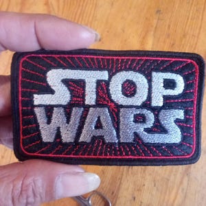 Puede incluir: Un parche bordado rectangular con fondo negro y un patr&oacute;n de rayos de sol rojos. El texto STOP WARS est&aacute; cosido con hilo blanco y plateado en una fuente negrita y estilizada. El parche presenta un borde negro grueso y esquinas redondeadas.