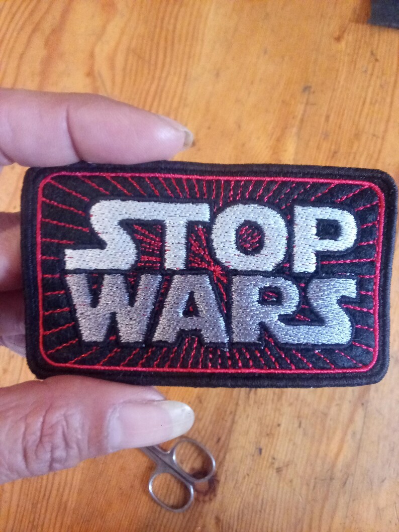 Puede incluir: Un parche bordado rectangular con fondo negro y un patr&oacute;n de rayos de sol rojos. El texto STOP WARS est&aacute; cosido con hilo blanco y plateado en una fuente negrita y estilizada. El parche presenta un borde negro grueso y esquinas redondeadas.