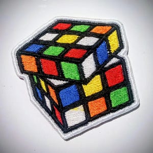 Könnte beinhalten: Gestickter Aufnäher mit einem bunten Rubik's Cube-Design. Der Aufnäher zeigt das klassische Puzzle mit seinen leuchtenden Quadraten in Rot, Orange, Gelb, Grün, Blau und Weiß, umrandet in Schwarz. Der Aufnäher hat einen weißen Rand.
