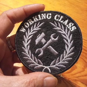Toppa rotonda nera con testo ricamato bianco "WORKING CLASS" e un'emblema di martello e chiave inglese. L'emblema è circondato da una corona d'alloro grigia.