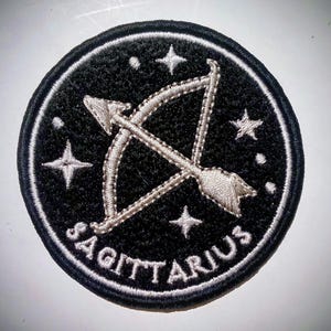 Può includere: Toppa rotonda ricamata nera con il simbolo zodiacale del Sagittario: un arco e una freccia. Il design include stelle ricamate bianche e la scritta "SAGITTARIUS" in lettere bianche. La toppa ha un bordo nero.