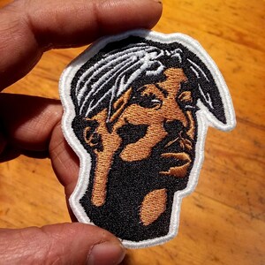 May include: Embroidered patch depicting a man with a black beard and hair, wearing a bandana. The patch has a white border and is held up by a hand. The portrait is in shades of black, brown, and white.