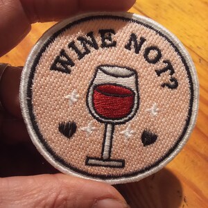 May include: Circular embroidered patch featuring the text "WINE NOT?" and a wine glass filled with red liquid. The patch is pale pink with a white border, black hearts, and white star accents.