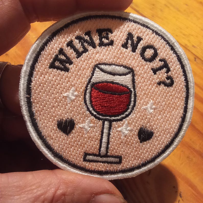 May include: Circular embroidered patch featuring the text "WINE NOT?" and a wine glass filled with red liquid. The patch is pale pink with a white border, black hearts, and white star accents.