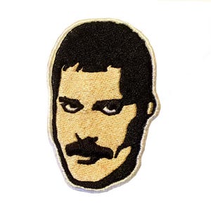 Op de afbeelding: Geborduurde patch met een gestileerd portret van een man met zwart haar en een snor. De patch heeft een bruin gezicht en zwart haar en snor, met een witte rand. Voor kleding of accessoires.