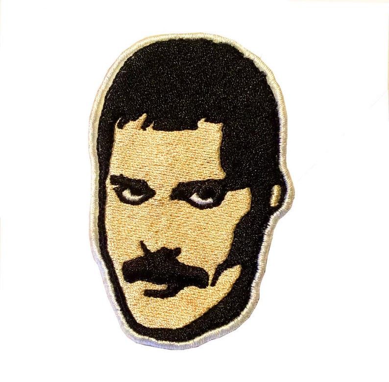 Op de afbeelding: Geborduurde patch met een gestileerd portret van een man met zwart haar en een snor. De patch heeft een bruin gezicht en zwart haar en snor, met een witte rand. Voor kleding of accessoires.