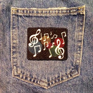 May include: A denim pocket with a black patch featuring embroidered musical notes in white, red, yellow, and green. The patch is rectangular and has a dark brown background. The pocket is part of a blue denim garment.