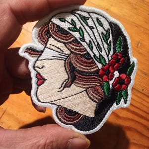 Może przedstawiać: Haftowana naszywka przedstawiająca profil kobiety z opaską na oczach, ozdobioną białą chustą i czerwonymi kwiatami. Naszywka ma czarną obw&oacute;dkę i jest wykonana z brązowych, czerwonych i zielonych nici. Około 7 cm wysokości.