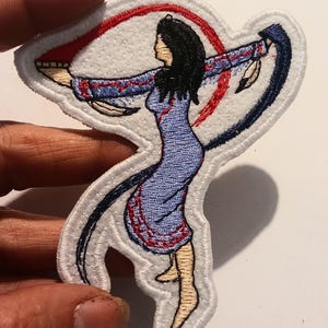 May include: Embroidered patch depicting a Native American woman in a blue dress with red and blue detailing. The patch has a white border and shows the woman with outstretched arms, encircled by red and blue curved lines.