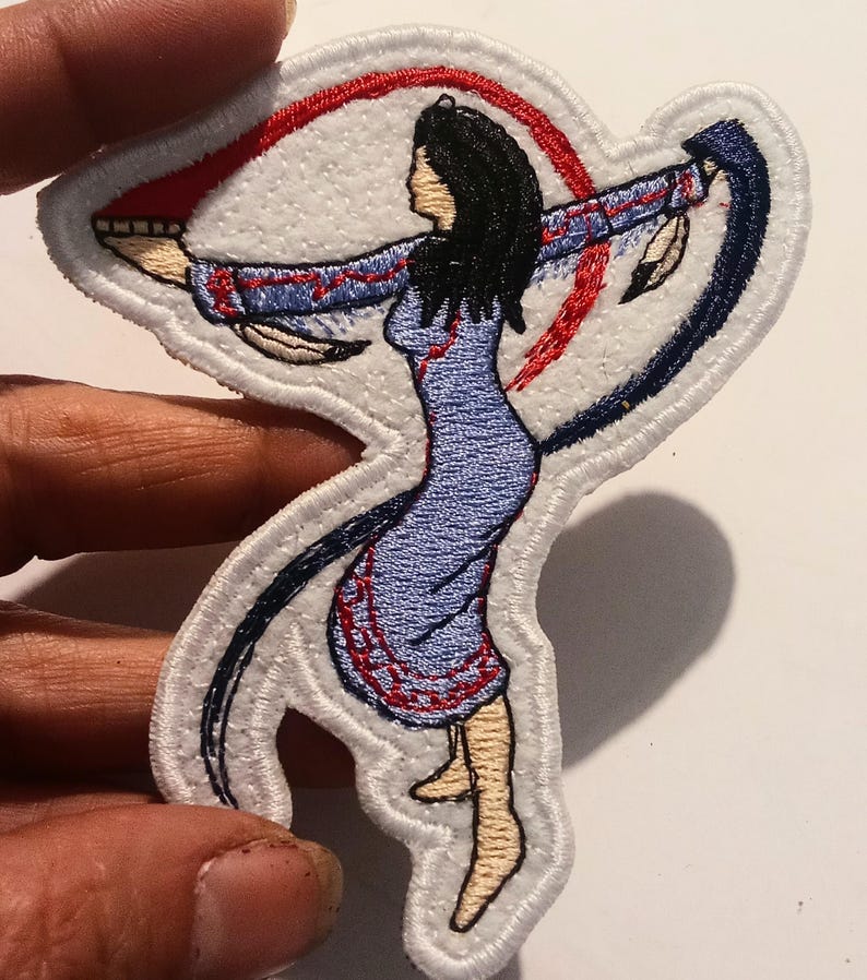 May include: Embroidered patch depicting a Native American woman in a blue dress with red and blue detailing. The patch has a white border and shows the woman with outstretched arms, encircled by red and blue curved lines.
