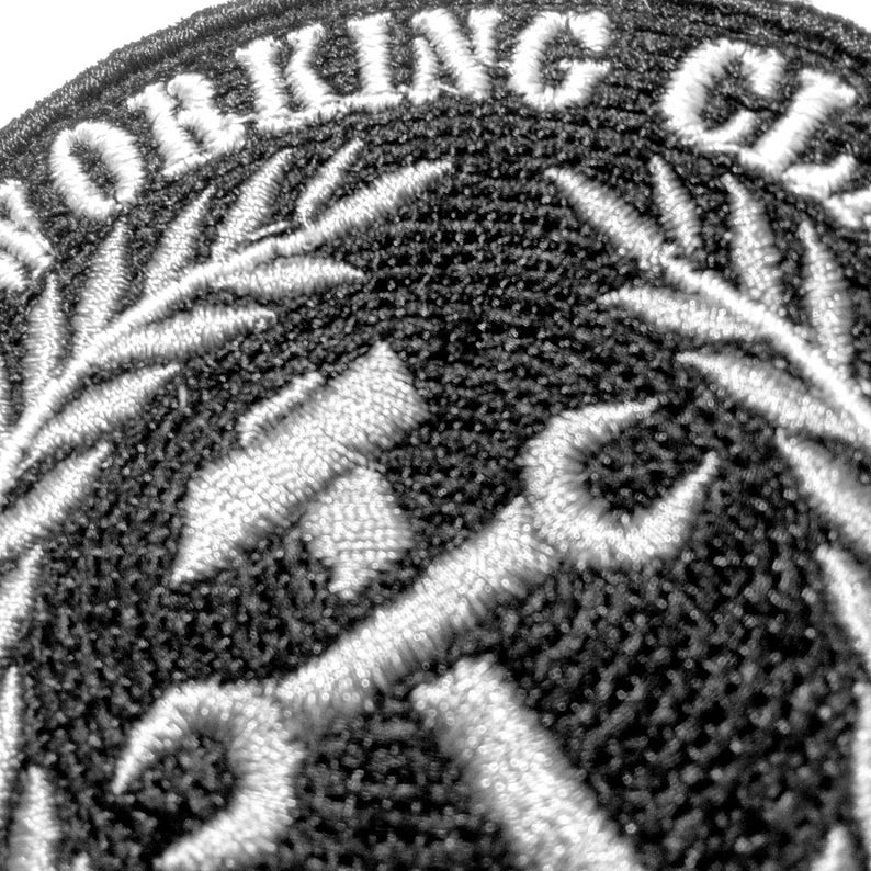 Toppa ricamata con la scritta "WORKING CLASS" in bianco, arcuata nella parte superiore. La toppa presenta un design a martello e chiave inglese al centro, circondato da una corona d'alloro bianca, il tutto su sfondo nero.