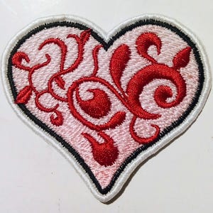 Può includere: Toppa ricamata a forma di cuore con sfondo rosa chiaro e design floreale rosso a spirale. La toppa ha un bordo nero e un bordo cucito bianco. Ideale per abbigliamento o accessori.