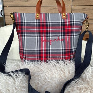 May include: A plaid tote bag in red, gray, black, and white with brown leather handles and a navy blue shoulder strap. The word "beijinho" is embroidered in red on the front of the bag. The bag has gold-colored hardware.