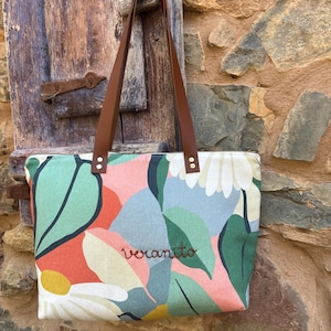 May include: A rectangular tote bag featuring a colorful botanical print with large leaves and flowers in shades of green, pink, blue, and yellow. The bag has brown leather straps attached with metal rivets and the word veranito embroidered in brown.