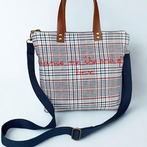 May include: A plaid tote bag with brown leather handles and a navy blue shoulder strap. The bag features red embroidered text that reads "Save me the end of love". The bag has a zipper closure.
