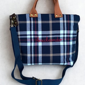 May include: A navy blue plaid tote bag with brown handles and a detachable blue shoulder strap. The bag features the embroidered word "bailando" in red. The bag has a rectangular shape.