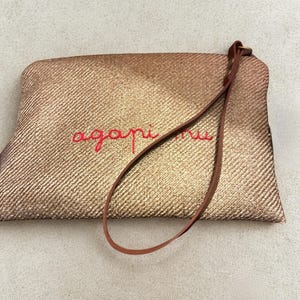May include: A gold-colored clutch with a woven texture. The word "agapi mu" is embroidered in red thread across the front. A brown leather strap is attached to the top of the clutch.