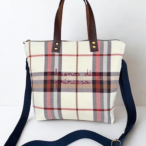 May include: Cream-colored tote bag with a plaid pattern in red, brown, and gray. It has brown leather handles and a navy blue shoulder strap. The text "buenos dias princesa" is embroidered in red.
