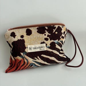 May include: A small, patterned pouch with a brown zipper and a wrist strap. The pouch features a mix of animal print designs, including cow spots, zebra stripes, and a floral pattern. The label reads "SAQUISAYO hecho a mano."