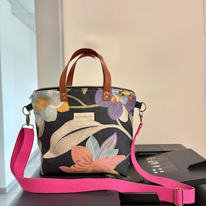 May include: A tote bag featuring a black fabric base with a colorful floral print in shades of orange, purple, and blue. It includes brown leather handles, a detachable bright pink woven shoulder strap, and a central label reading SAQUISAYO.