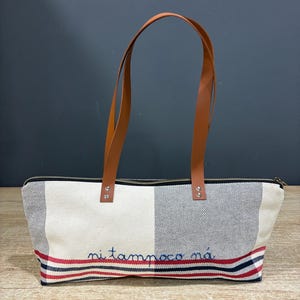 May include: A rectangular tote bag featuring a cream, navy blue, and red plaid pattern. The bag has two long tan leather straps attached with silver-tone metal rivets. The text "ni tampoco ná" is embroidered in blue on the front lower section.