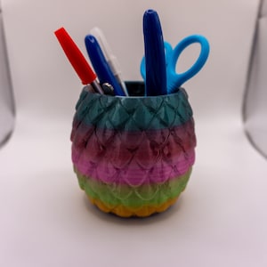 May include: A multi-coloured pen holder with a scale-like design, holding a red pen, a white pen, a blue pen, and blue scissors. The holder transitions from green to pink to yellow.