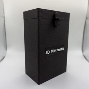 May include: A black rectangular box with a textured surface and a hinged lid. The box has a small rectangular label with the text "3D Materials" in white. The box is set against a white background.