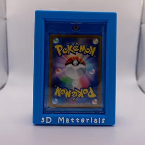 May include: A blue 3D-printed frame containing a Pokémon trading card. The card displays the Pokémon logo and a colourful illustration. The frame has the text "3D Materials" at the bottom.