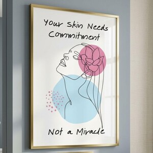 May include: A framed art print with a gold frame, featuring a line drawing of a woman's face looking upwards. The artwork includes a pink flower and blue circle, with the text "Your Skin Needs Commitment, Not a Miracle."