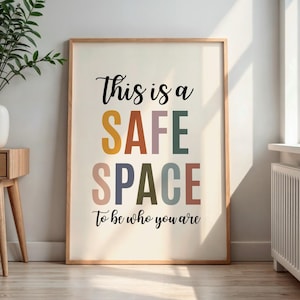 Pode incluir: Uma impressão emoldurada com as palavras "This is a SAFE SPACE to be who you are" em uma fonte colorida e divertida. As letras são em várias cores, incluindo amarelo, roxo, verde e marrom. A moldura é de cor de madeira clara.