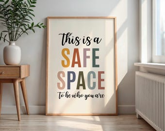May include: A framed print with the words "This is a SAFE SPACE to be who you are" in a colorful, playful font. The letters are in various colors, including yellow, purple, green, and brown. The frame is a light wood color.