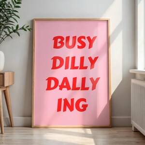May include: A framed pink art print with the words "BUSY DILLY DALLY ING" in bold red letters. The artwork is displayed in a light-colored wooden frame, leaning against a white wall. A side table with a vase of greenery is to the left.