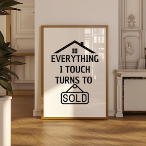 May include: A framed print featuring the text "EVERYTHING I TOUCH TURNS TO SOLD" in black. The design includes a house silhouette and a "SOLD" sign. The frame is gold, and the background is white.